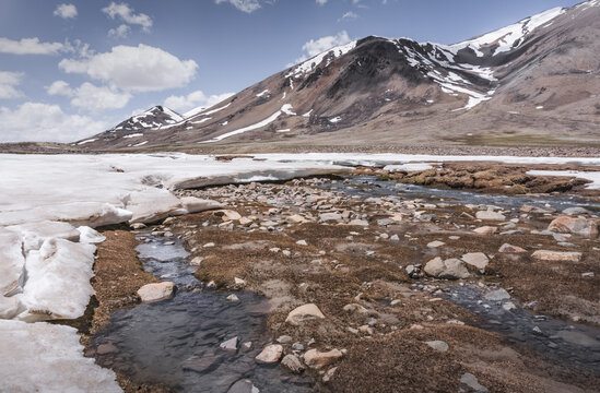 Landscape in the high Pamirs with rocky peaks and a river in a valley with melting snow. Panorama of rocky mountains on a sunny day and glaciers in a river valley