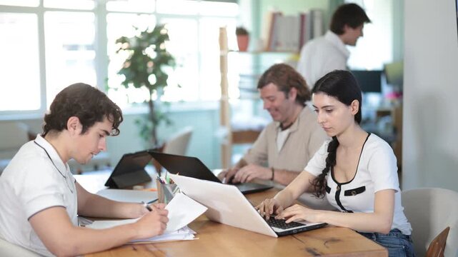  Male and female colleagues sit at desks using laptops and reviewing paper documents in office