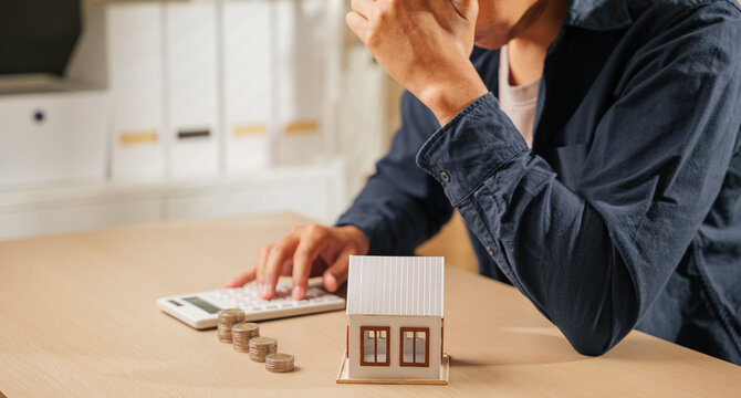 Stressed person holding a calculator, with stacks of coins and a house model nearby. Reflects financial planning challenges, tax concerns, and property calculations causing worry