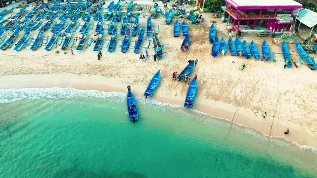 Overhead aerial view shows rows of blue fishing boats spread across pale sand beside turquoise water at Ngerenean Beach.