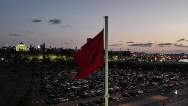 Blue hour drone view of the Moroccan flag overlooking Hassan Tower, the V Mausoleum and visitor facilities in Rabat, showcasing Morocco's historic capital and heritage tourism.