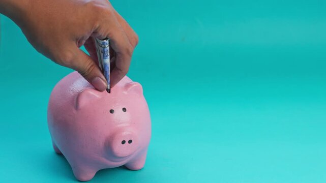 Hand puts paper money into pink piggy bank on turquoise background