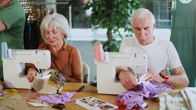 Man and woman focusing on stitching at machines during group class for seniors in tailoring studio, while fellow learners preparing pattern pieces for assembly and adjusting fabric on mannequin