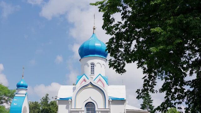 Static shot of ornate blue dome and golden cross of Saints Boris and Gleb Orthodox Cathedral framed by green tree branches against cloudy summer sky in Daugavpils Latvia.