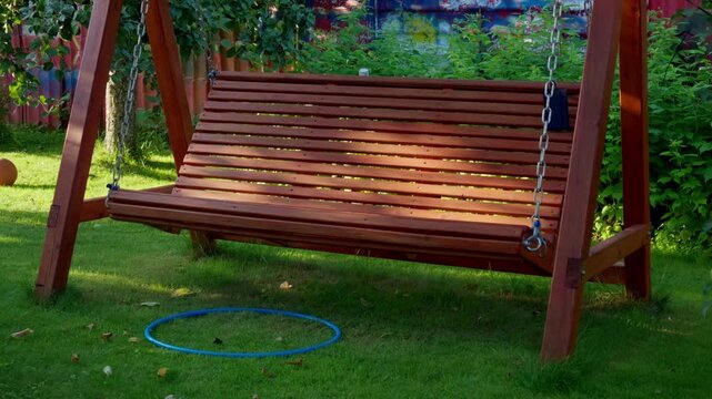 Empty brown wooden bench swing hangs on chains over grassy ground with plastic toy hoop. Summer afternoon scene in peaceful domestic yard surrounded by green foliage.