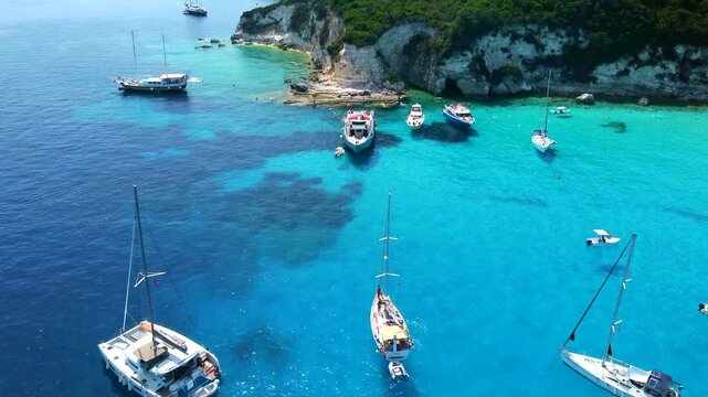 Aerial fly-in toward the beautiful rocky shores of the Greek island of Antipaxos, showcasing luxury yachts and swimmers enjoying the vibrant turquoise Ionian Sea
