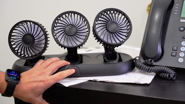 Close-up of a person's hand pressing a red power button on a three-head desktop cooling fan. This small electronic device is used to improve air circulation at an office desk workstation.