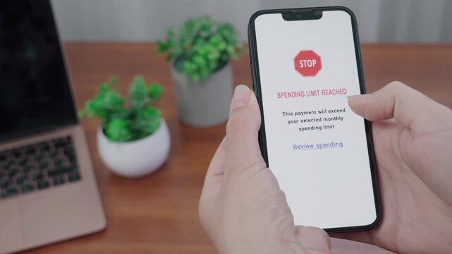 Person holds a phone displaying a red STOP spending limit reached message from an AI finance tool over a wooden desk with laptop and plants.