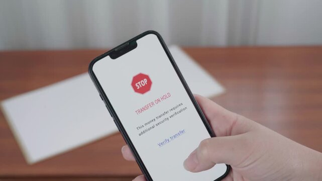 Hand holds a mobile device with an AI security message that a money transfer is on hold pending extra verification.