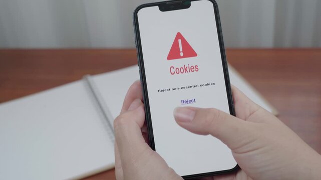 User holds a smartphone displaying a cookie consent warning screen and moves a thumb toward the reject non essential cookies option.