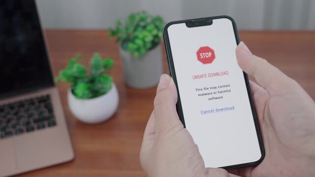 User holds a phone displaying an AI information unsafe download warning and hovers over the cancel download link at a wooden table.