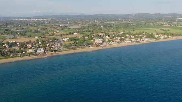 Kourouta Beach in Ilia, Greece, showing a long sandy shoreline, calm blue sea, a small coastal settlement, and agricultural land stretching inland