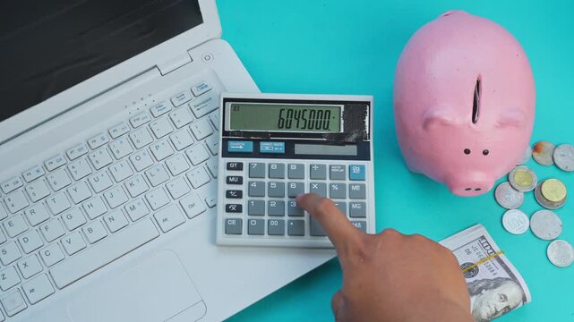 Hand uses calculator next to laptop, piggy bank, cash, and coins on turquoise background