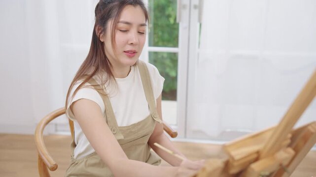 Happy Asian woman artist in beige dungarees sitting and painting watercolor with paintbrush on canvas on wooden easel in clean white bright home studio with calm, relaxation and concentration