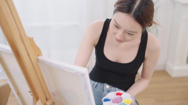 Beautiful, happy Asian Japanese woman artist in black tank top painting watercolor with paintbrush on canvas on wooden easel in clean white bright home studio room with calm, relaxation and happiness