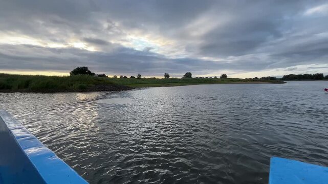 Wide River Landscape Viewed from Moving Vehicle Ferry During Summer River Crossing with Calm Water, Green Shoreline, Cloudy Sky and Natural Riverside Scenery
