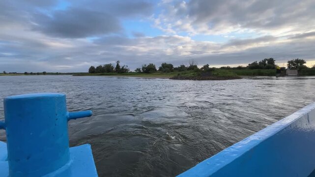 Ferry Wake and Flowing Water Viewed from Moving Vehicle Ferry During Summer River Crossing with Calm River Surface, Cloudy Sky and Riverside Landscape