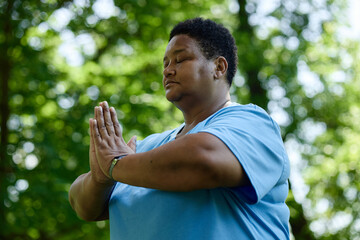 Naklejka premium Middle aged African American woman meditating with closed eyes and hands in prayer position in park, promoting mental wellbeing and outdoor wellness campaigns, medium shot