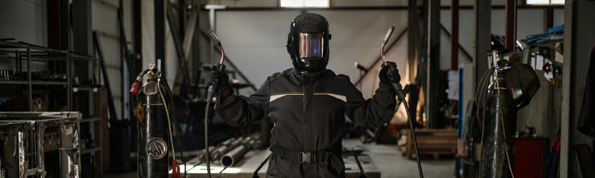Portrait of welder in protective mask holding welding torches in industrial workshop. Image represents manufacturing expertise and is suitable for metalworking industry marketing