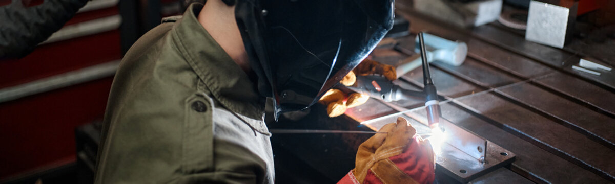 Young adult Caucasian male welder in protective mask welding metal with colleague in workshop, representing teamwork. Image suitable for industrial training and manufacturing marketing