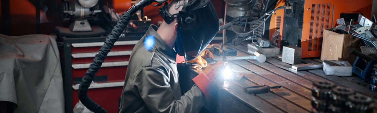 Caucasian young adult male welder working in industrial workshop, welding metal parts. Image suitable for vocational training programs and manufacturing industry marketing, wide shot