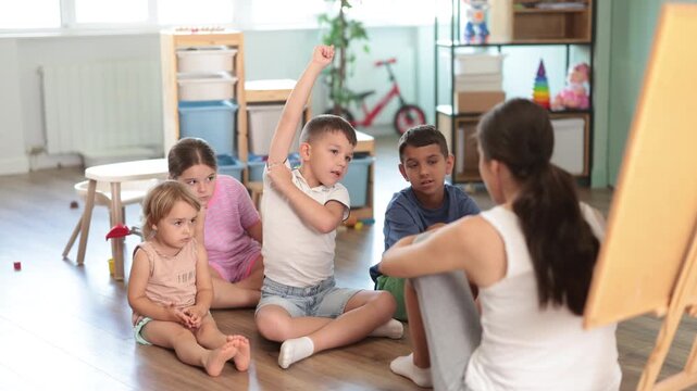 Glad diverse kids paying attention to young mentor during lesson in cozy kindergarten classroom