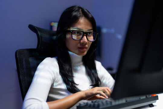 Asian woman with glasses working at computer monitoring system data and cybersecurity dashboard at night, focused analyst watching screen in dark office