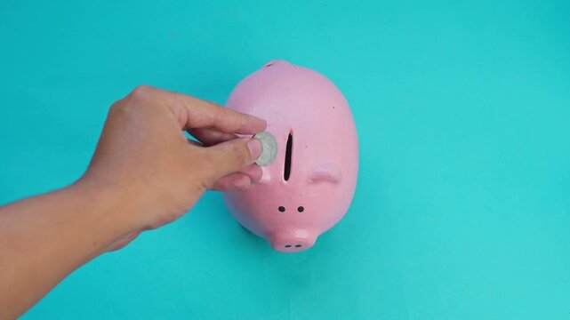 Hand drops coin into pink piggy bank on turquoise background