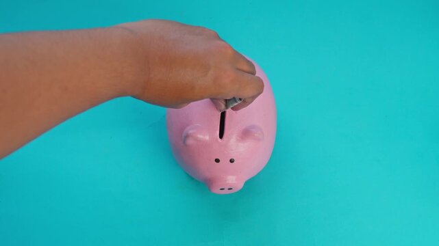 Hand inserts cash into pink piggy bank on turquoise background