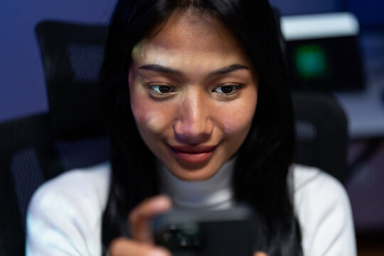 Close up of a young woman smiling as she looks down at her smartphone in a dark room lit by blue and green screen glow, absorbed in a mobile app at night.