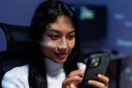 Woman holds a smartphone and taps the screen while sitting at a desk at night, code and data glowing on monitors behind her, suggesting online shopping or mobile banking.
