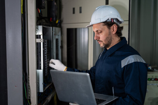 Professional male electrical engineer in uniform using laptop computer for inspecting control panel and switchboard in smart factory. Industrial maintenance and automation technology concept.