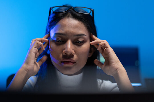 Close up of a woman with glasses pushed up rubbing her temples and staring at a bright monitor in a blue room, showing eye strain from long screen hours.