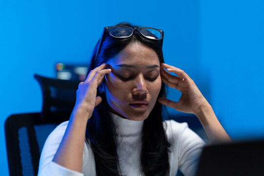 Woman developer closes her eyes and presses her temples with glasses pushed up, sitting at a laptop in a blue lit room during late night programming work.