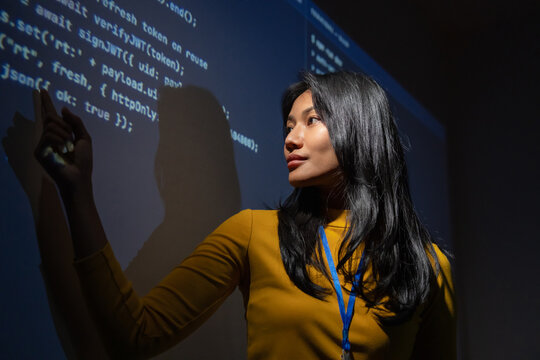 Woman technology professional reaches toward projected code in a dark room, hand raised as if interacting with a virtual interface, suggesting AI and digital transformation.
