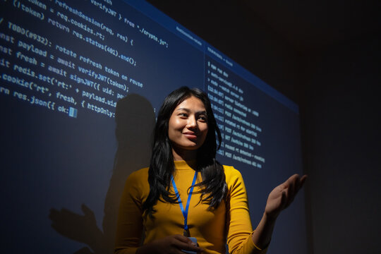 Woman developer smiles with an open hand gesture while answering a question, backend code and server logs projected behind her during a team review session.