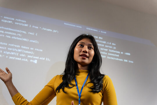 Young woman in yellow top with lanyard listens during a coding seminar, hand raised mid conversation, programming code projected on the screen behind her.