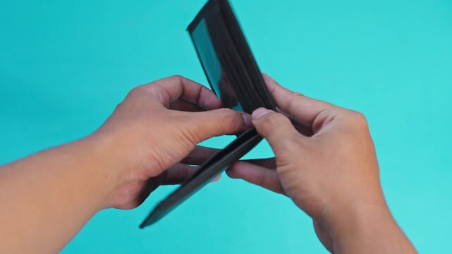 Hands open empty black wallet letting remaining coins fall out onto turquoise background