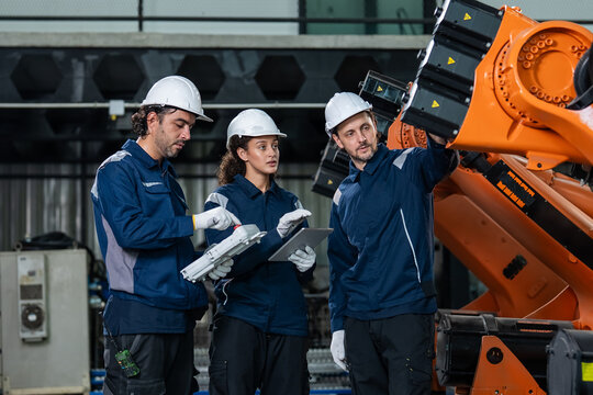 Diverse engineering team in blue uniforms using a digital tablet to program an industrial robotic arm in a modern smart factory, highlighting Industry 4.0 automation and collaboration.