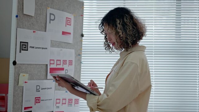 young female designer stands beside a pinboard filled with branding sketches and logo drafts in a bright, modern office, thoughtfully analyzing design variations for a corporate identity project.
