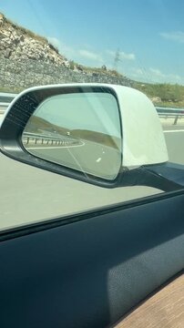 Car side mirror reflection of open scenic highway road and sunny mountain landscape while driving in EV electric vehicle on summer road trip