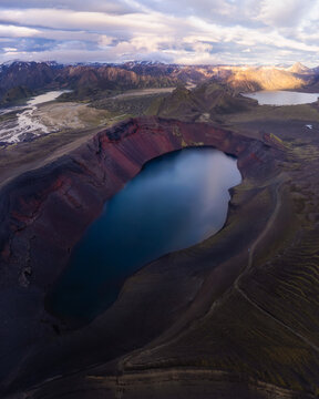 Aerial view of Ljotipollur crater lake with its striking red volcanic slopes and deep blue water surrounded by rugged mountains under a dramatic cloudy sky in Iceland.