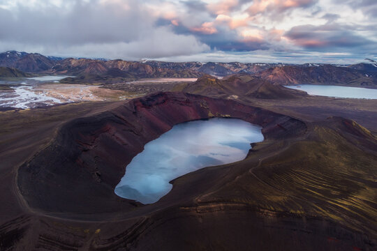 Aerial view of Ljotipollur volcanic crater lake with vibrant red slopes and dark basaltic rock formations set against a dramatic cloudy sky in Iceland.