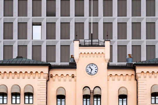 Centered beneath a grid, the clock crowns an ornate facade in Oslo, Norway, where historic symmetry meets modern architecture as windows reinforce the contrast.