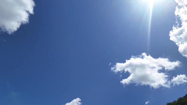 Cielo azzurro con nuvole bianche sparse e sole brillante sopra una linea di alberi verdi. Scena luminosa, naturale e serena.