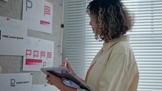 young female designer stands beside a pinboard filled with branding sketches and logo drafts in a bright, modern office, thoughtfully analyzing design variations for a corporate identity project.
