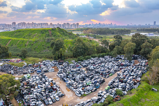 Aerial view of a car scrapyard filled with rows of wrecked vehicles, near a green hill with an urban skyline in the background at sunset in Petah Tikva, Center District, Israel.