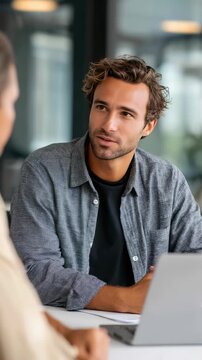 A focused young man communicates attentively with a coworker in a bright office environment. He is using a laptop and sharing insights in a collaborative work atmosphere