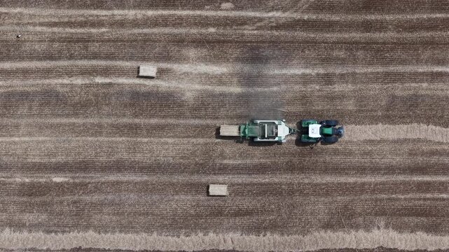 An aerial view captures a modern tractor and baler working in a vast, harvested field, efficiently processing straw into bales.