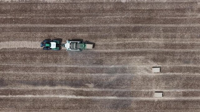 A tractor and baler efficiently produce rectangular hay bales in a vast harvested field, an aerial view illustrating modern agricultural practices.